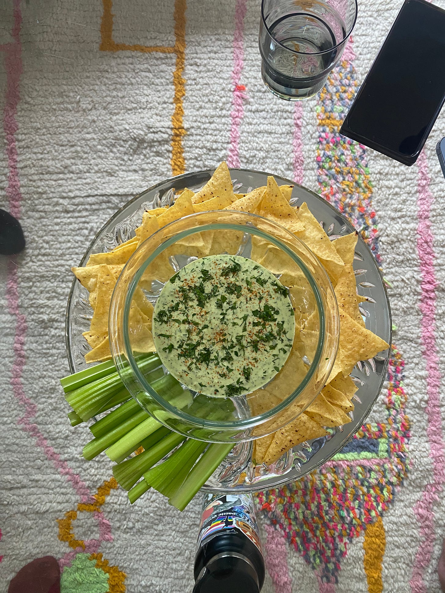 A glass plate with a glass bowl set in the centre, on top of a glass coffee table: the plate has tortilla chips and celery sticks, and the bowl has the green dip, dusted with herbs and smoked paprika.