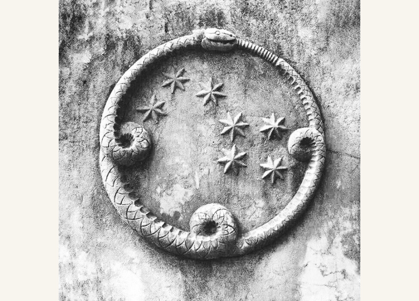 Black and white photo of the ouroboros symbol, a snake eating its own tail. In the centre of the circle formed by the snake are spreaded seven 6-pointed stars. This whole piece is sculpted on a wall.
