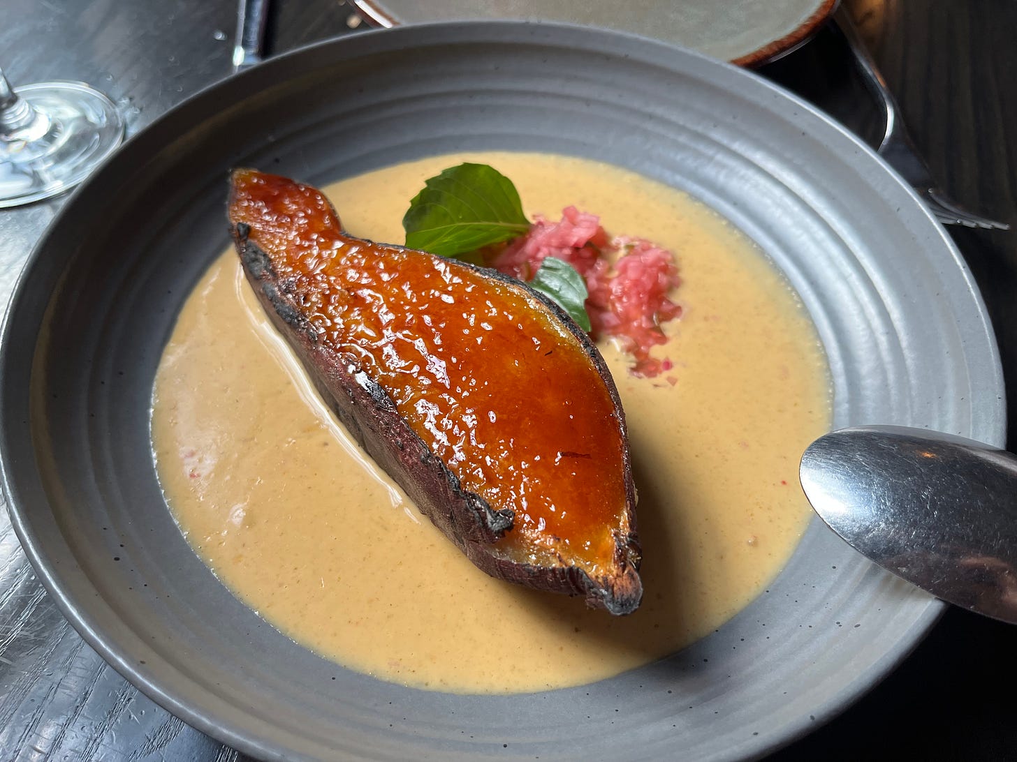 a long half of a sweet potato with skin on, cut side up, with the surface covered in red-brown caramelized sugar, basil leaves, and pink pickled food that has been minced, all on a bed of yellow-orange curry, on a round gray plate