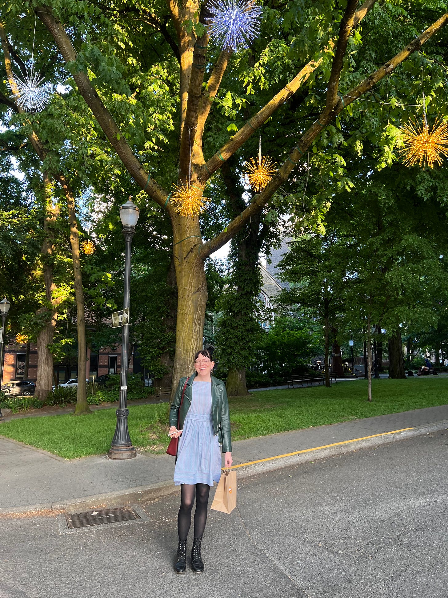 Emme stands in a blue dress and tights under a tree in the park blocks in downtown portland. Twinkling light globes hang in the tree above her. The light is golden. Emme stands in a blue dress and tights under a tree in the park blocks in downtown portland. Twinkling light globes hang in the tree above her. The light is golden.