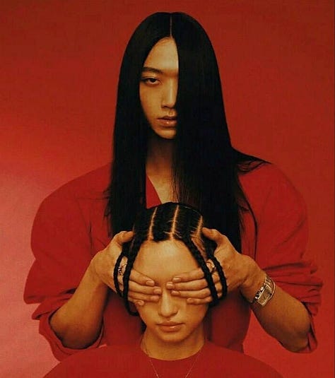 Three stylized studio portraits against vivid orange-red backgrounds: on the left, a person with long black hair covers another person’s eyes from behind; in the center, a person with red rope wrapped around their eyes and mouth; on the right, a person crouches while a high-heeled shoe presses against their open mouth, with a small pink bottle on the floor nearby.