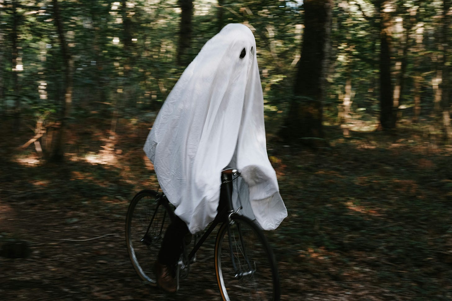 a person on a bicycle in a forest who is wearing a sheet with eyes cut out to resemble a ghost