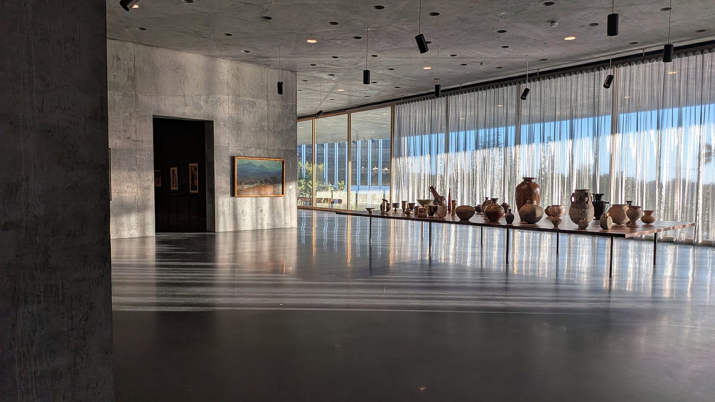 A wide view inside the Geffen Galleries showing floor-to-ceiling drapes filtering daylight along a curved glass wall, with concrete columns and an open, minimalist gallery floor. A wide view inside the Geffen Galleries showing floor-to-ceiling drapes filtering daylight along a curved glass wall, with concrete columns and an open, minimalist gallery floor.