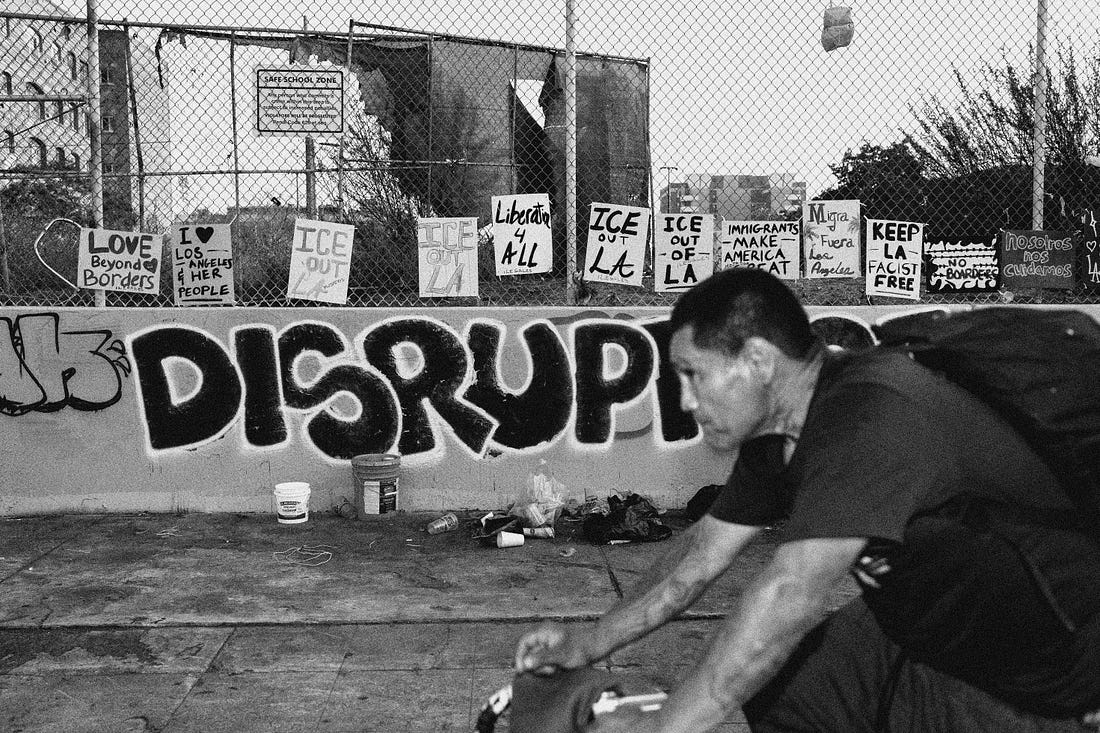 A man rides a bike past a wall with graffiti that says DISRUPT and that is lined with anti-ICE protest signs like "ICE out of LA" and "Love Beyond Borders"