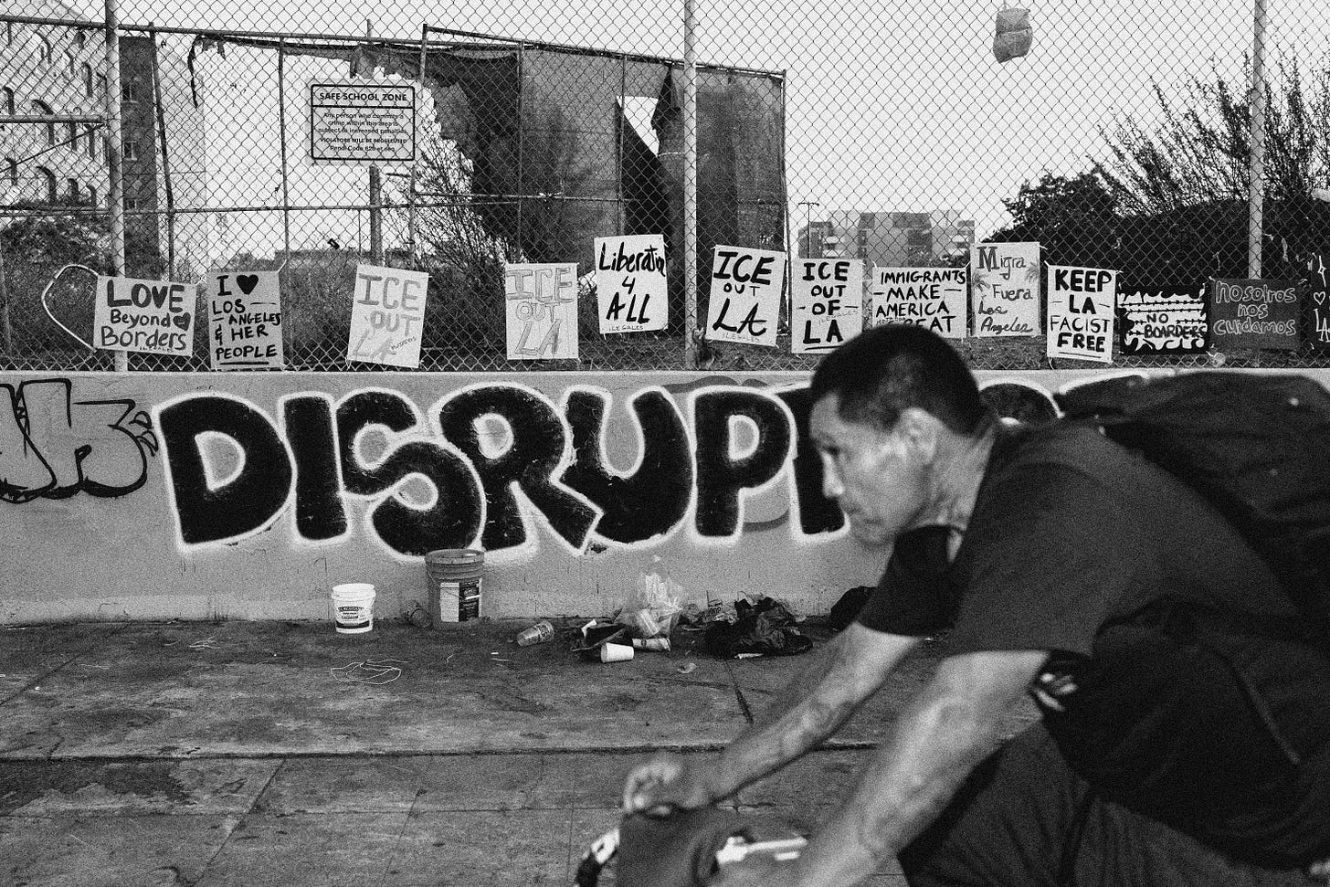 A man rides a bike past a wall with graffiti that says DISRUPT and that is lined with anti-ICE protest signs like "ICE out of LA" and "Love Beyond Borders" A man rides a bike past a wall with graffiti that says DISRUPT and that is lined with anti-ICE protest signs like "ICE out of LA" and "Love Beyond Borders"