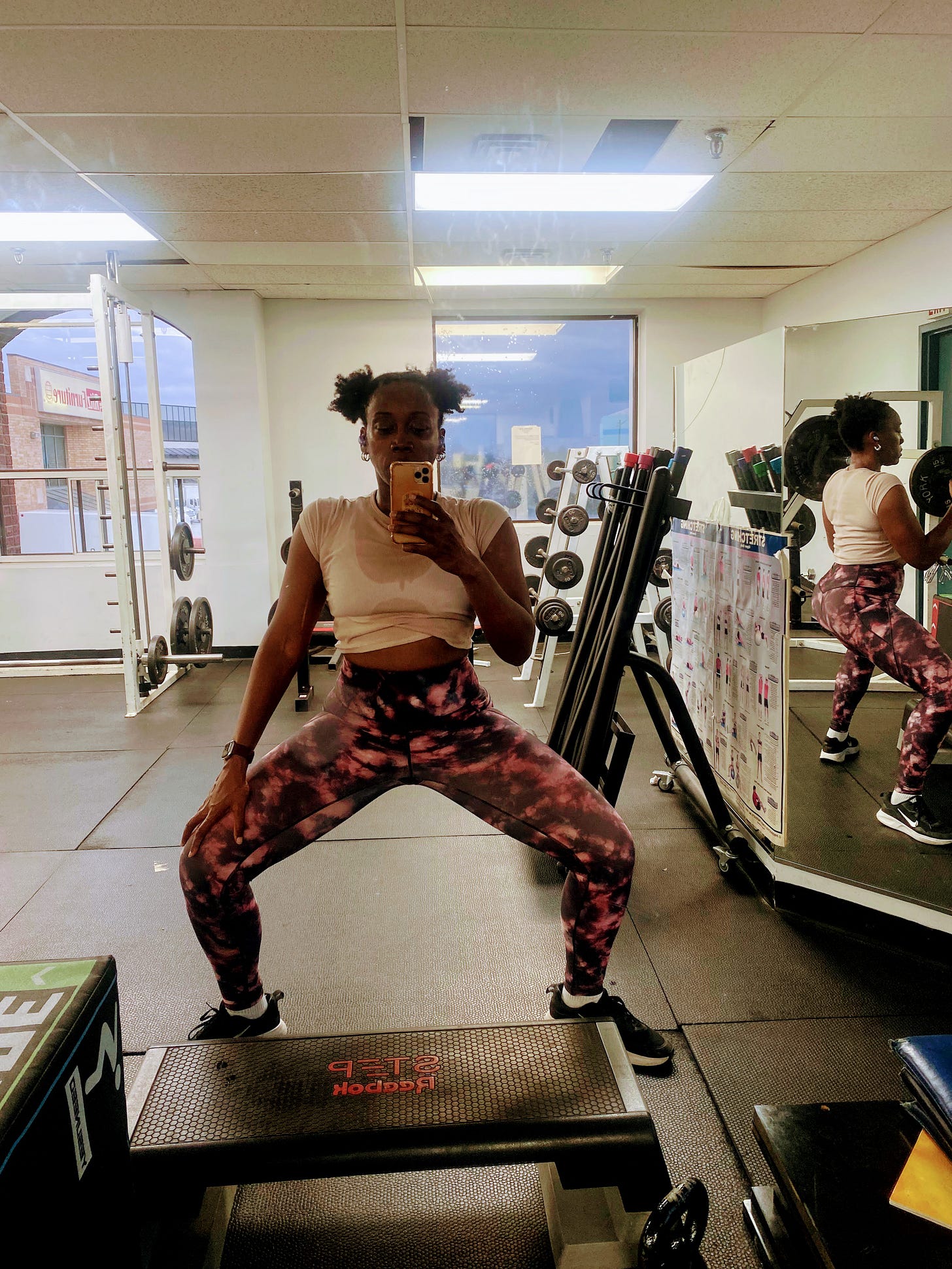 a sweaty woman at a gym with hair in afropuffs standing in a plié squat taking a selfie.