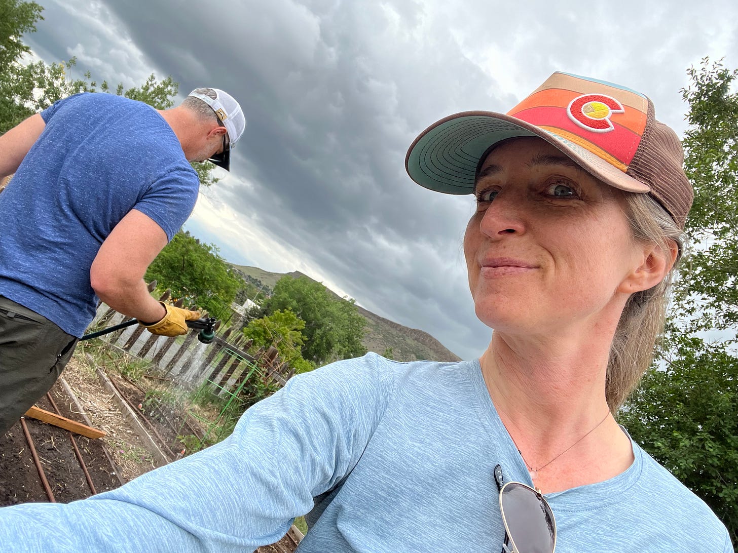 A photo of the author wearing a ball cap and smiling at the camera while her person sits over her shoulder facing away watering the garden A photo of the author wearing a ball cap and smiling at the camera while her person sits over her shoulder facing away watering the garden