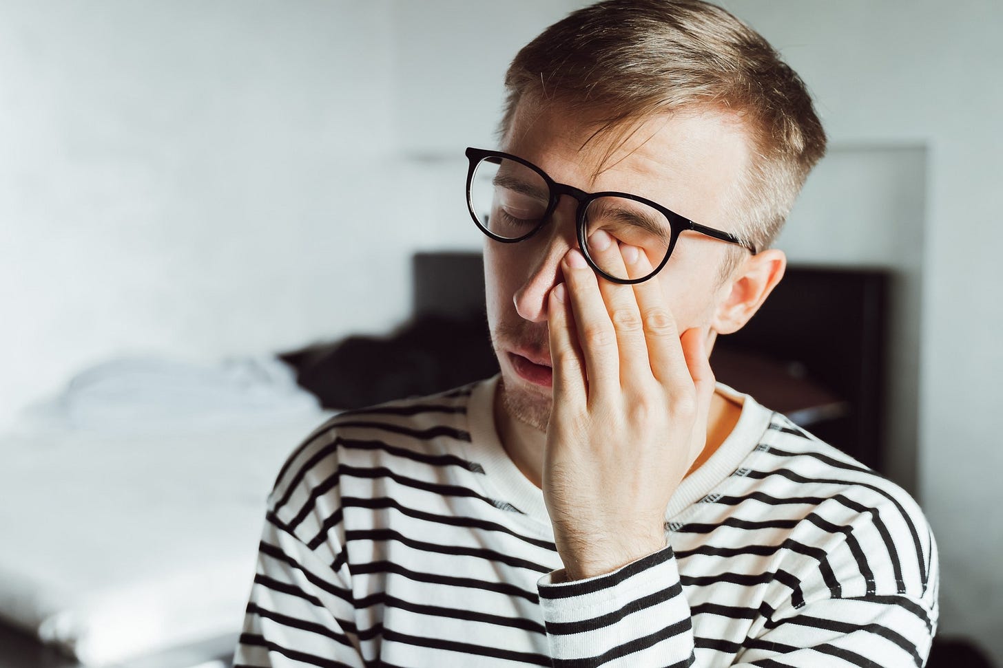 A man in a striped shirt sighs and rubs his face. His finger slide beneath his glasses.