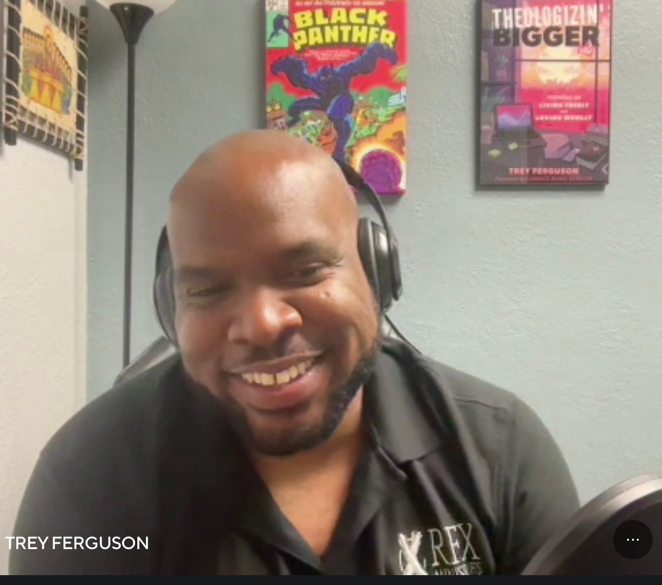 Pastor Trey, a Black man with a beard smiling and wearing headphones, sitting at a desk with a podcast mic. Behind him there is a Black Panther comic wall hanging as well as one of the cover of his book, Theologizin' Bigger