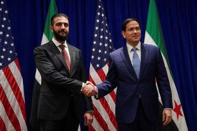 Syrian President Al-Sharaa and a US general shaking hands. They are dressed in suits, standing in front of American and Syrian flags on a blue curtain backdrop.