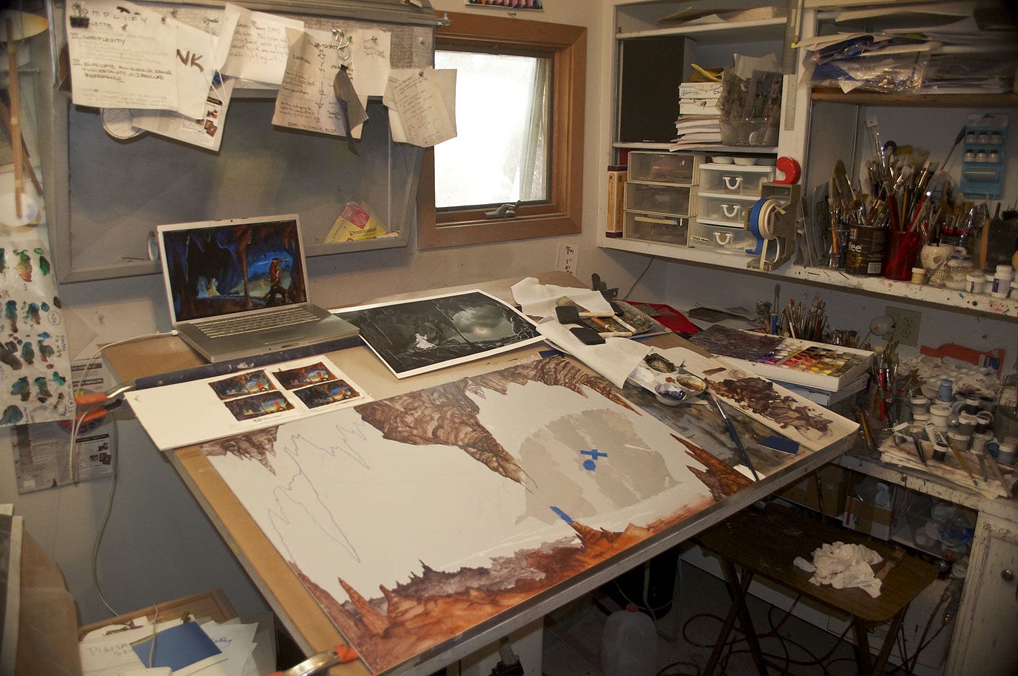 Photo of the illustration space in the artist's studio with A MEMORY OF LIGHT as a work in progress on the drafting table. The immediate foreground is being rendered with cave background area still white and the cave opening gray. At the top of the angled drafting table sits a laptop computer. To its right is a large black and white preliminary concept. In front to the left is a series of four small color studies set unevenly on white paper.