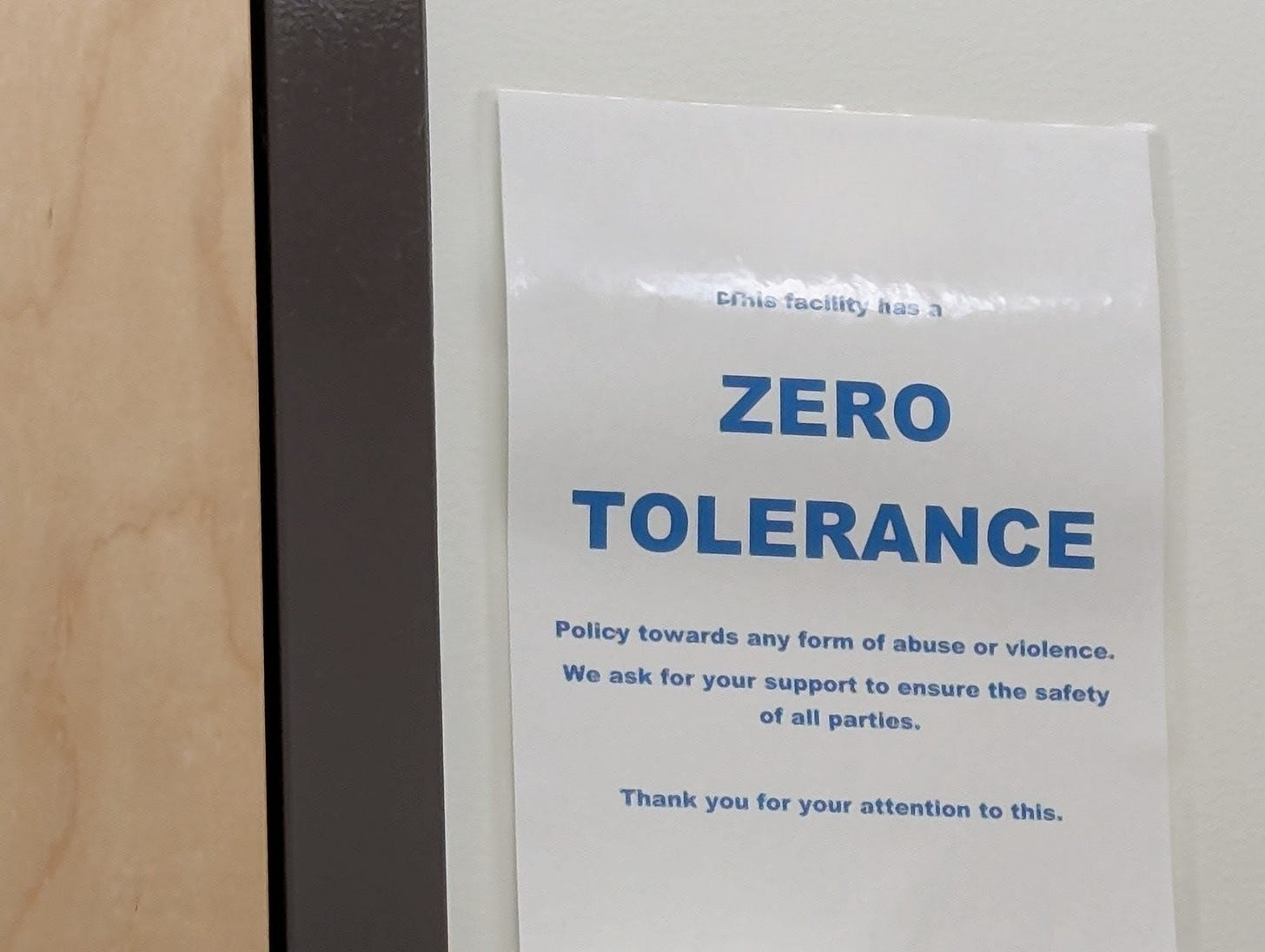 A white sign on the wall of a business with blue lettering that says, “This facility has a zero tolerance policy towards any form of abuse or violence.” A white sign on the wall of a business with blue lettering that says, “This facility has a zero tolerance policy towards any form of abuse or violence.”