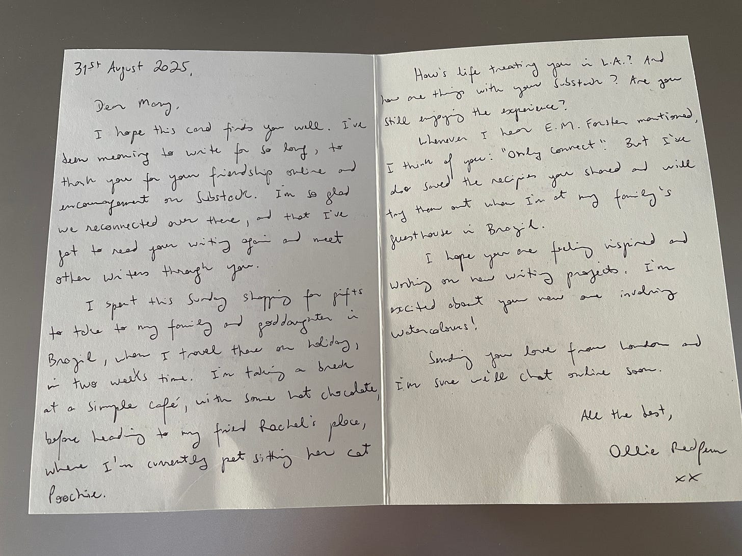 A handwritten card displayed on a cafe table. The card shows Ollie's handwriting in blue ink on cream paper, dated 31st August 2025, asking Mary about her life and Substack while mentioning his upcoming trip to Brazil. The card sits next to another envelope on a light wooden surface.