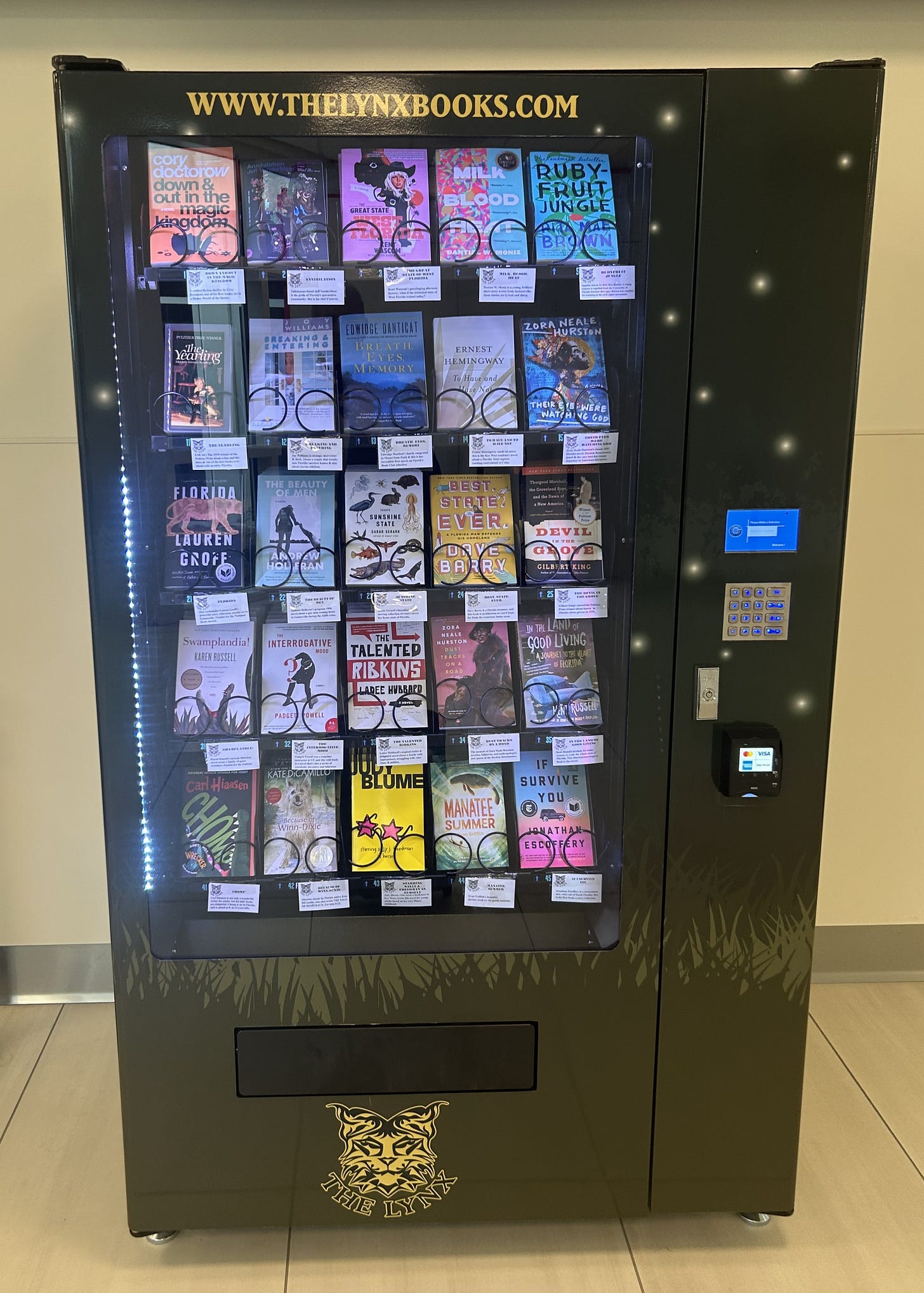 A black vending machine positioned against a white wall. It has a website address at the top an an image of a lynx at the bottom. It contains twenty-five, five books each in five rows.