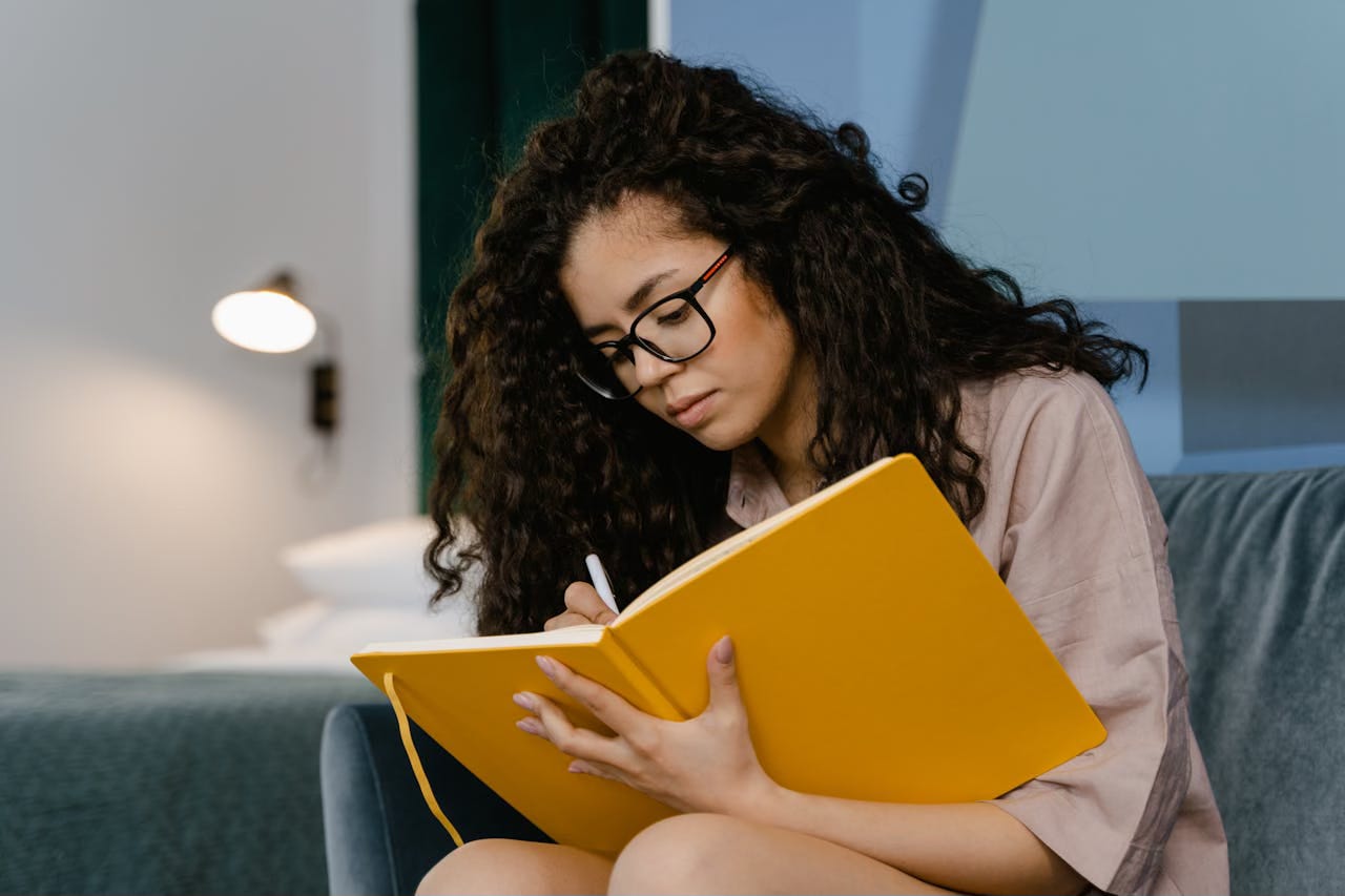 A woman with long, dark curly hair sits in an armchair writing in a journal with a bright gold cover.