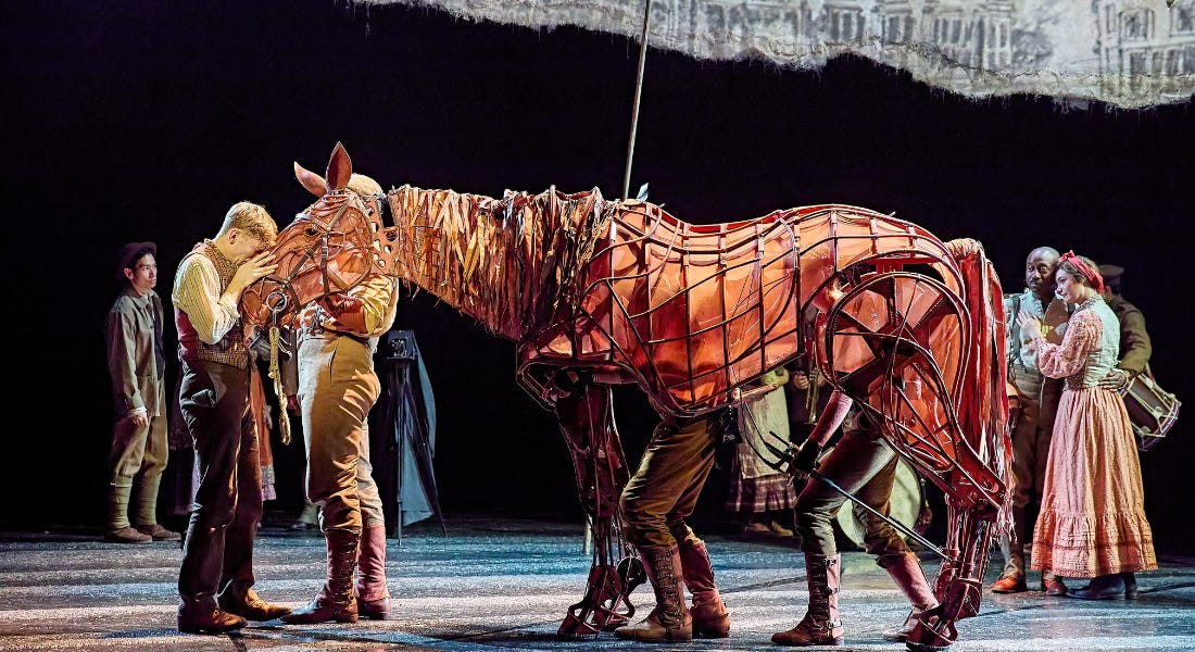 A theatre stage with a man next to a brown puppet horse, with actors and a black screen behind him