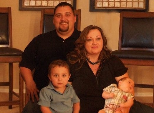 Posed family portrait with dad with toddler sitting in front of him and mom holding a baby who refuses to look at the camera. Mom and dad are wearing black and the kids light blue and light colored plaid.  Everybody but the baby looks mildly amused.