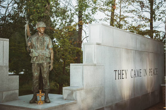 Bronze statue of a U.S. Marine standing with rifle before a granite memorial wall inscribed “THEY CAME IN PEACE,” surrounded by trees at the Beirut Memorial in Jacksonville, North Carolina, honoring the 241 Americans killed in the 1983 Marine Barracks bombing.