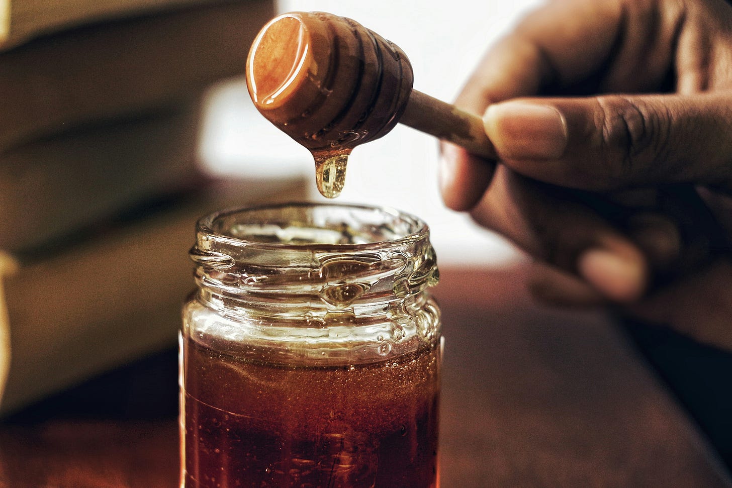 Hand holding a honey dipper with honey dripping into a glass jar.