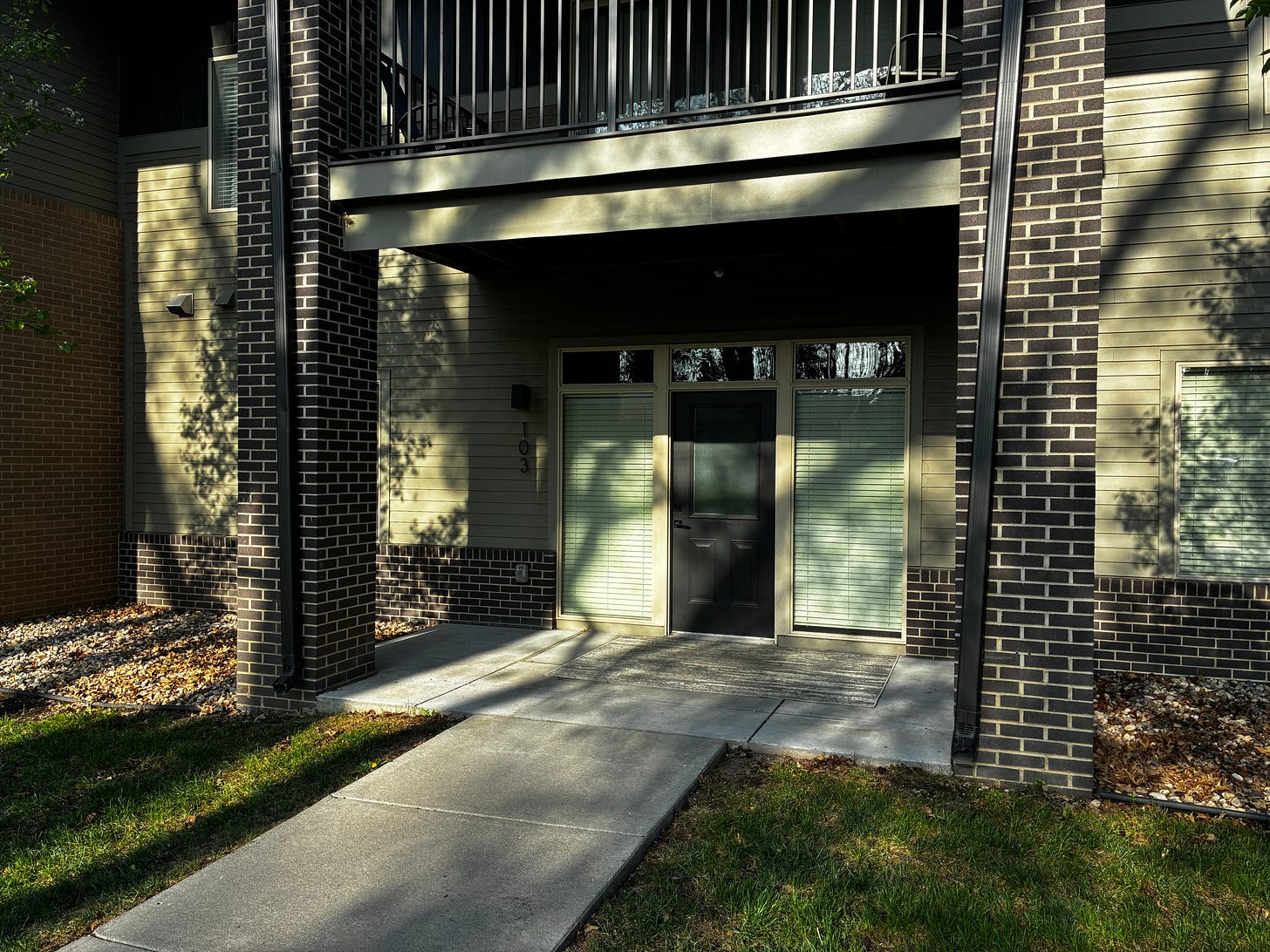 A small patio in dappled sunlight with a dark green entrance door flanked by two large windows with mini blinds