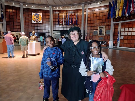 Annette with a lifesize mural of President Gerald Ford; Pulling a small covered cart on two wheels; with two kids in a museum atrium; With staues of Franklin and Eleanor Roosevelt sitting at tables; taking picture of photo exhibit; Outdoors with monuments and road in background.
