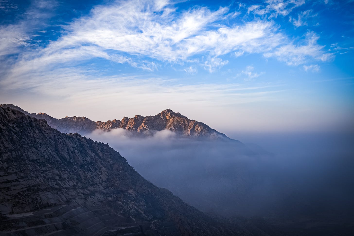 A dramatic view of rugged mountain peaks in Al Hada, Saudi Arabia, partially obscured by a thick layer of white mist and clouds under a bright blue sky.