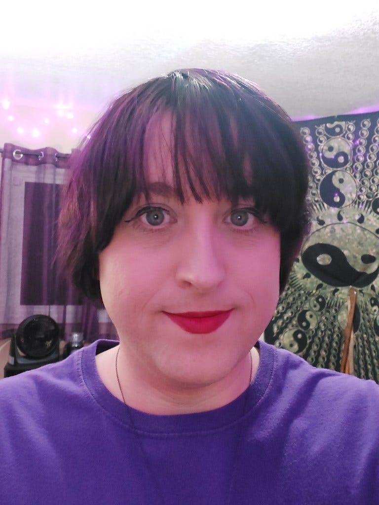 This image is a selfie of Lily, a person with short, dark hair and bangs, smiling slightly while looking at the camera. She is wearing a purple shirt and red lipstick. The background features a tapestry with yin-yang symbols and string lights.