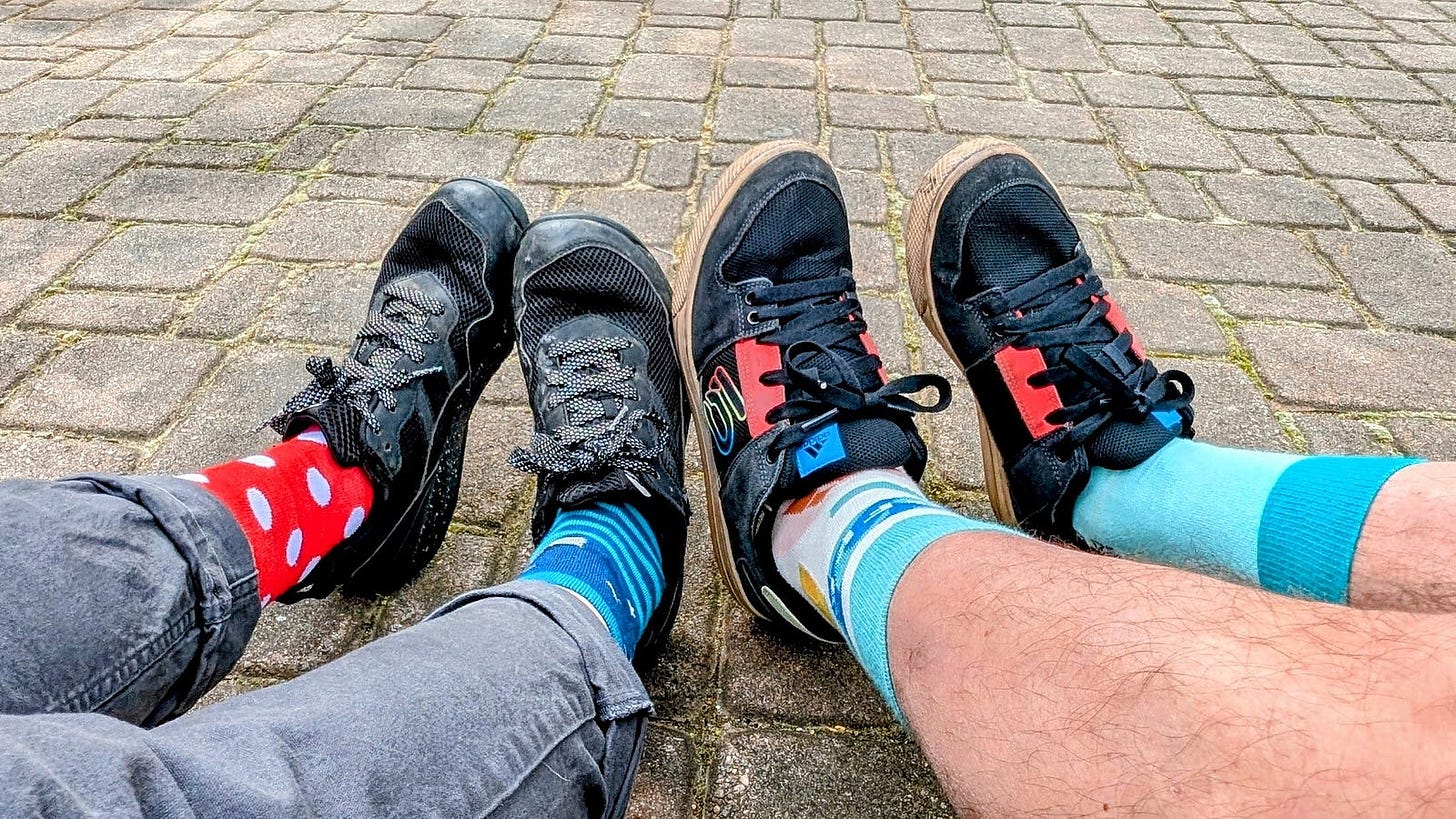 The author and Abby sit side by side on a brick patio, feet forward in black flat-soled sneakers. Each wears mismatched socks for 321 Day: red with white polka dots and blue stripes on the left, white patterned and light blue on the right. The author and Abby sit side by side on a brick patio, feet forward in black flat-soled sneakers. Each wears mismatched socks for 321 Day: red with white polka dots and blue stripes on the left, white patterned and light blue on the right.