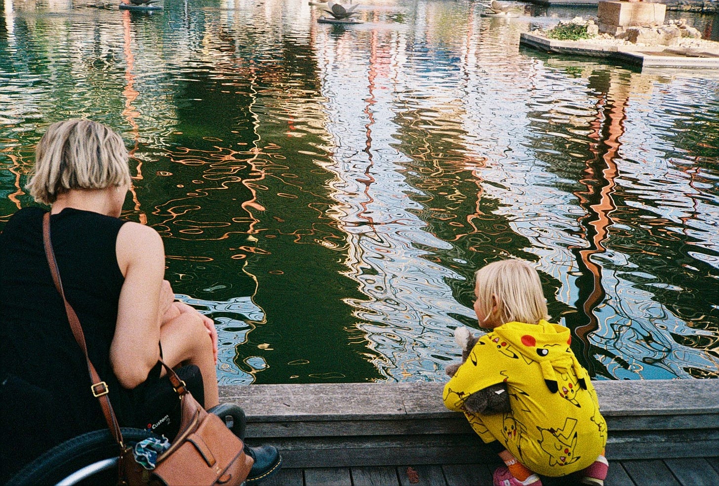Rebekah and her son face a glossy pond with ripple-waves that zigzag and pool like a wet painting. Otto crouches down, close to the water. Rebekah sits in her wheelchair close by. They're looking in the same direction.