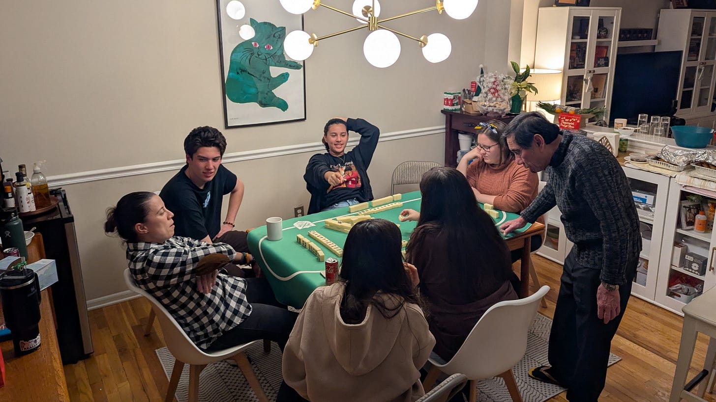 Dining room table covered in felt for a mahjong game. There are mahjong tiles on the table, some built into four walls in front of each player along with the tiles that each player is using. Additional chairs surround the table for observers. The body language is leaned back, relaxed, happy. An older person stands besides the table and is teaching the young players how to play mahjong