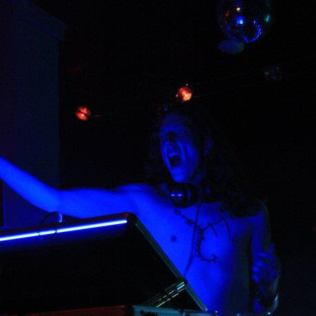 A picture of Satanic Fragment, shirtless and bathed in blue light, screaming with his arm out stretched while DJing