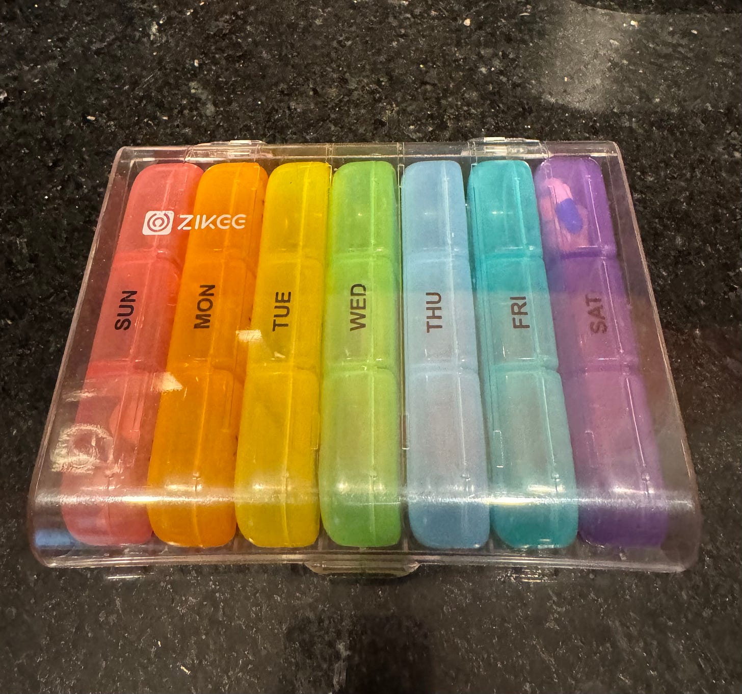 A clear, plastic container holding seven rainbow-colored pill organizers.