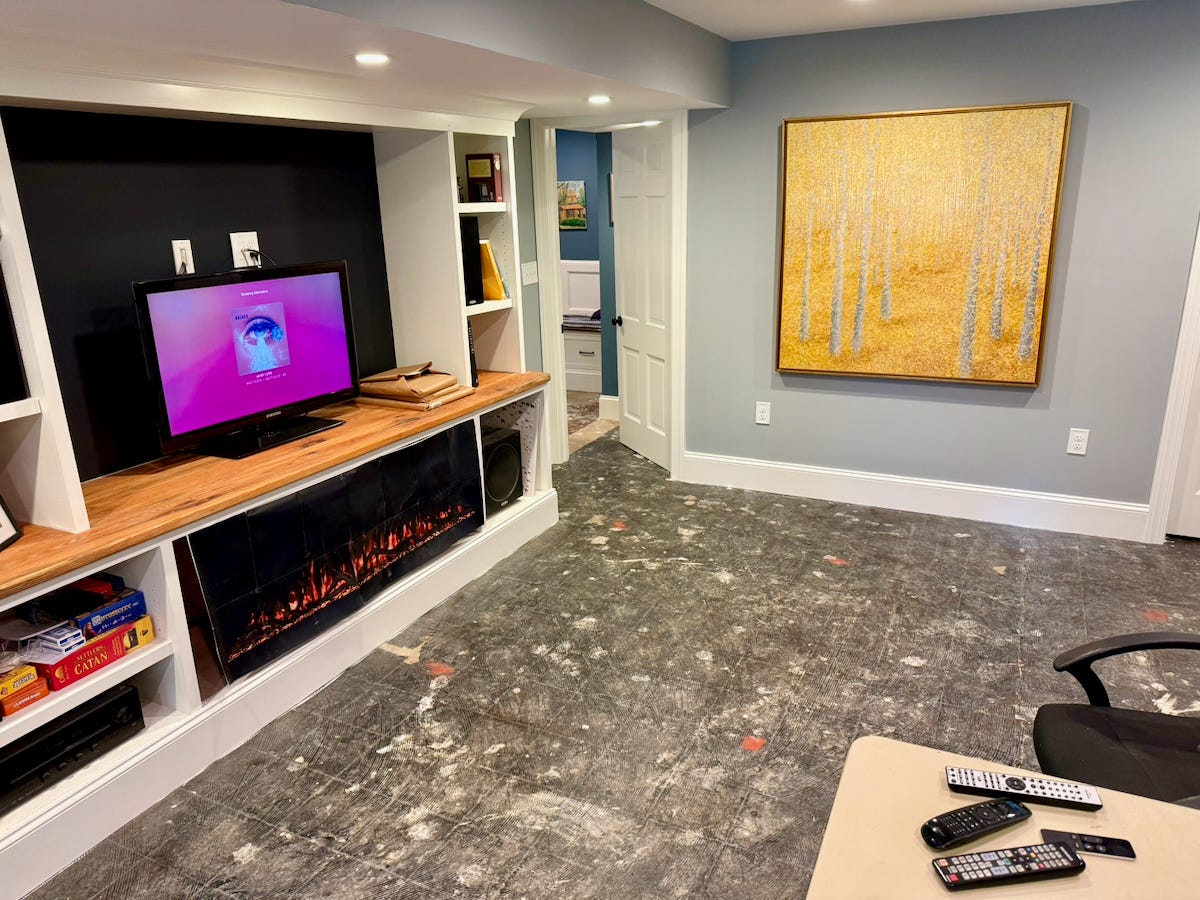 Basement with built-in shelving, a television, and an electric fireplace beside a bare concrete floor mottled with paint, joint compound, and old adhesive residue.
