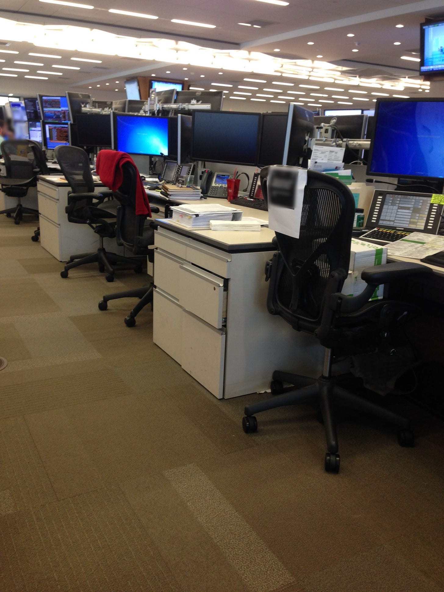 A busy investment-bank trading floor with rows of desks, walls of computer monitors, and empty office chairs under bright fluorescent lights. A busy investment-bank trading floor with rows of desks, walls of computer monitors, and empty office chairs under bright fluorescent lights.