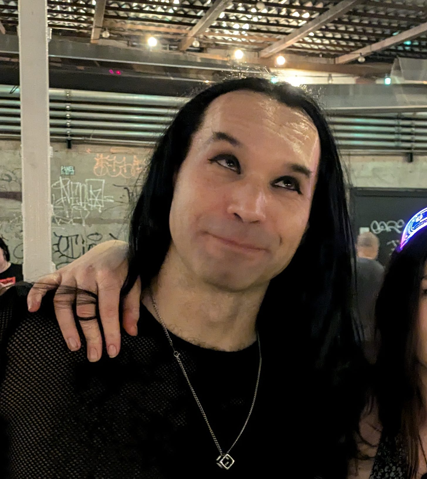 Person with long dark hair wearing black shirt and geometric pendant necklace.