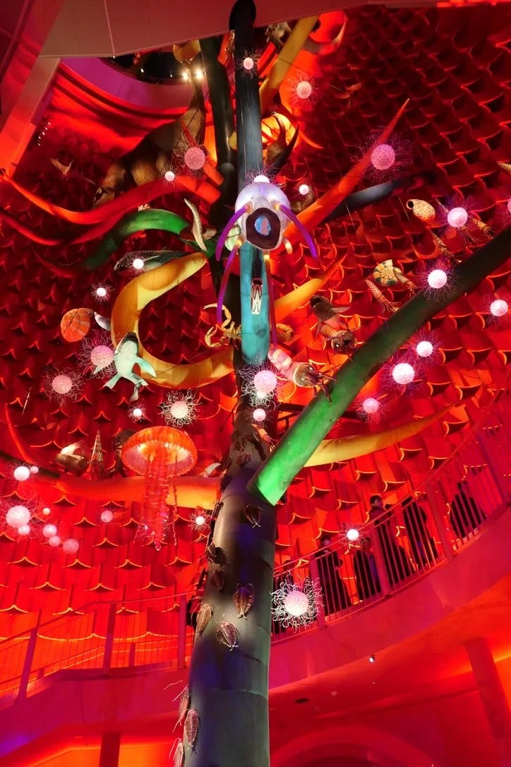 A colorful sculpture of a tree, with models of animals attached to different branches, looked at from below. The sculpture is in a cavernous space with red lighting