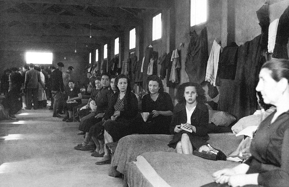 #descrevemospravocê  Fotografia em preto e branco, de época, feita em um alojamento coletivo, com dezenas de pessoas reunidas em um ambiente interno amplo e pouco iluminado. A cena mostra principalmente mulheres sentadas lado a lado sobre camas ou colchões simples, dispostos em fileira ao longo das paredes. Elas vestem roupas escuras, com expressões sérias ou cansadas, e algumas seguram objetos nas mãos, como xícaras, pratos ou pedaços de pão. À frente, uma mulher aparece em destaque, virada de perfil, com as mãos entrelaçadas.  O alojamento tem teto alto com estrutura de madeira aparente, paredes lisas e janelas retangulares posicionadas na parte superior. Nas paredes laterais, estão pendurados cobertores, roupas e toalhas. Ao fundo, à esquerda da imagem, um grupo de pessoas — homens e mulheres — está de pé, formando uma pequena aglomeração, conversando ou aguardando algo. O piso é liso e de cimento.  A imagem transmite uma atmosfera de espera, incerteza e sobriedade, sugerindo um cenário de deslocamento forçado, como um campo de refugiados, centro de acolhimento ou abrigo durante uma guerra ou crise humanitária.