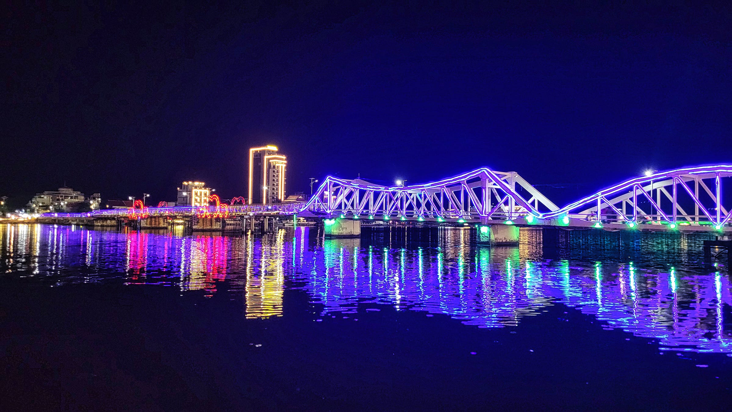 Illuminated bridge in Kampot, Cambodia, at night, with colorful lights reflecting on the river.