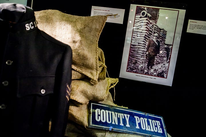 Photographs showing tin hats with 'Police' written on them, bullseye lamps, a hand bell, metal County Police signs and medals.