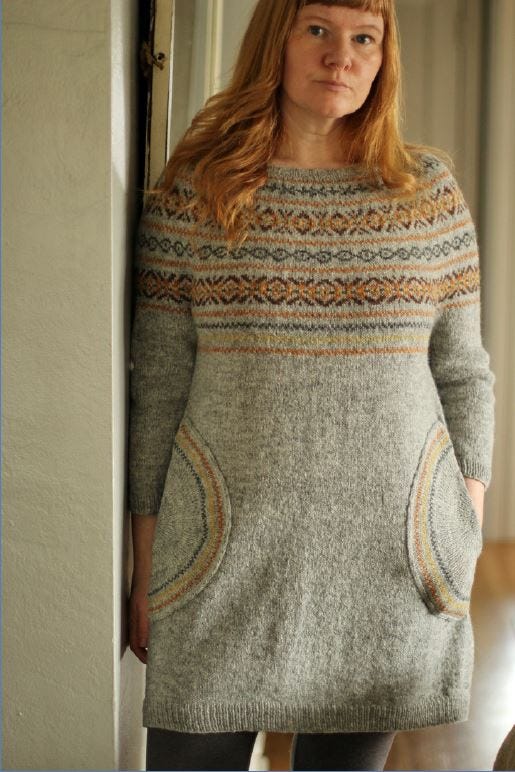 A woman with red hair stands in the doorway looking at the camera, wearing a long grey knitted tunic with Fair Isle details around the collar and pockets A woman with red hair stands in the doorway looking at the camera, wearing a long grey knitted tunic with Fair Isle details around the collar and pockets