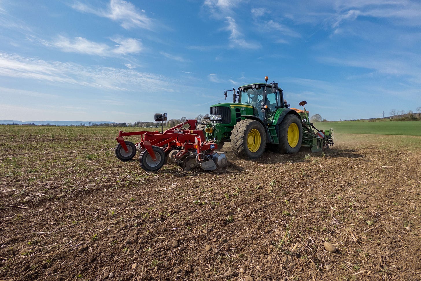 Ein grüner John Deere-Traktor fährt über ein Feld. An der Front ist eine Scheibenegge montiert, hinten eine Kreiselegge kombiniert mit einer pneumatischen Sämaschine. Die Maschinenkombination dient der flachen Bodenbearbeitung und gleichzeitigen Aussaat. Im Hintergrund erstreckt sich eine sanft hügelige Landschaft unter blauem Himmel.