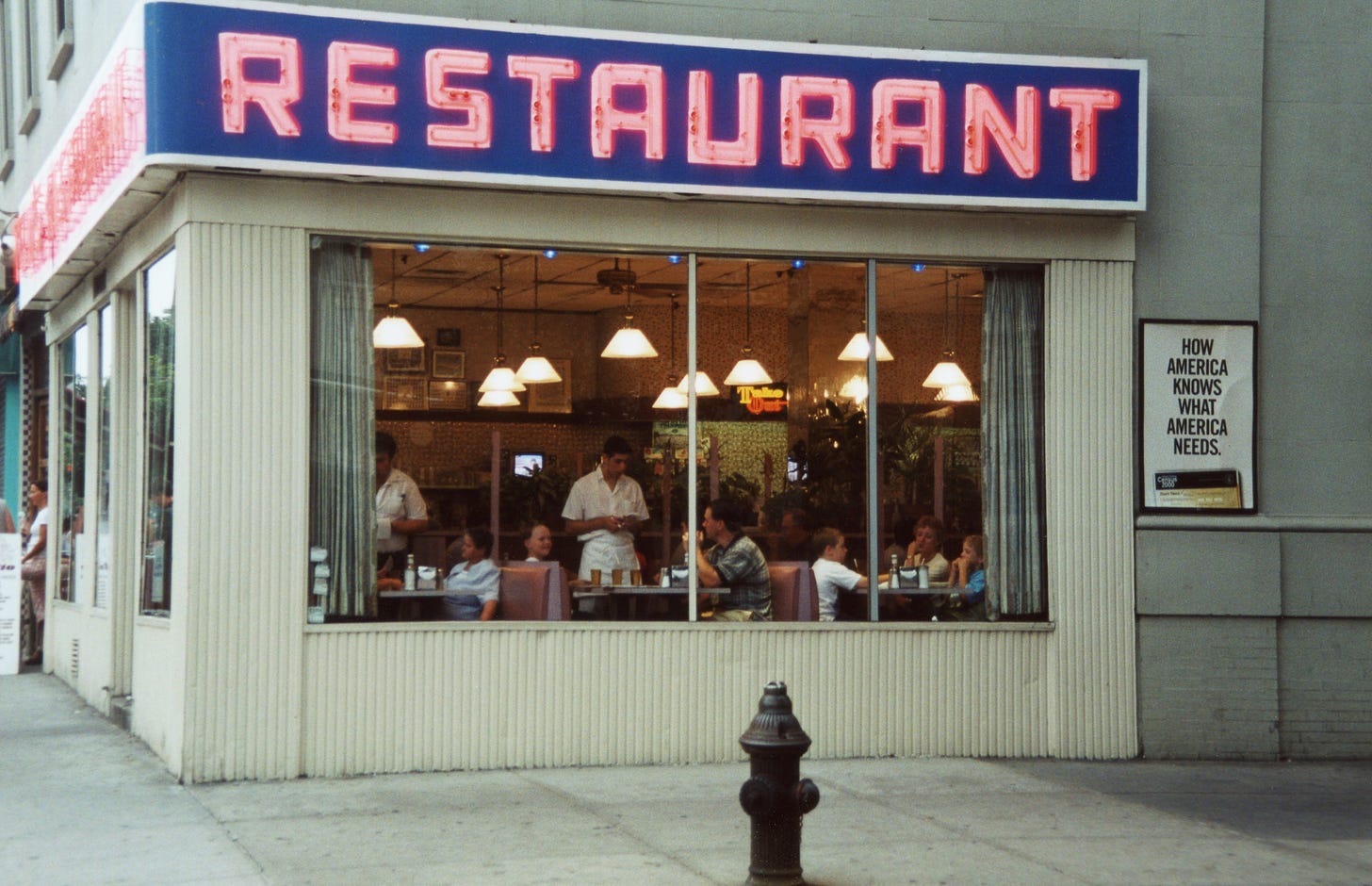A corner diner with a blue sign reading "RESTAURANT" in red letters. Pitcure windows show servers taking orders inside.