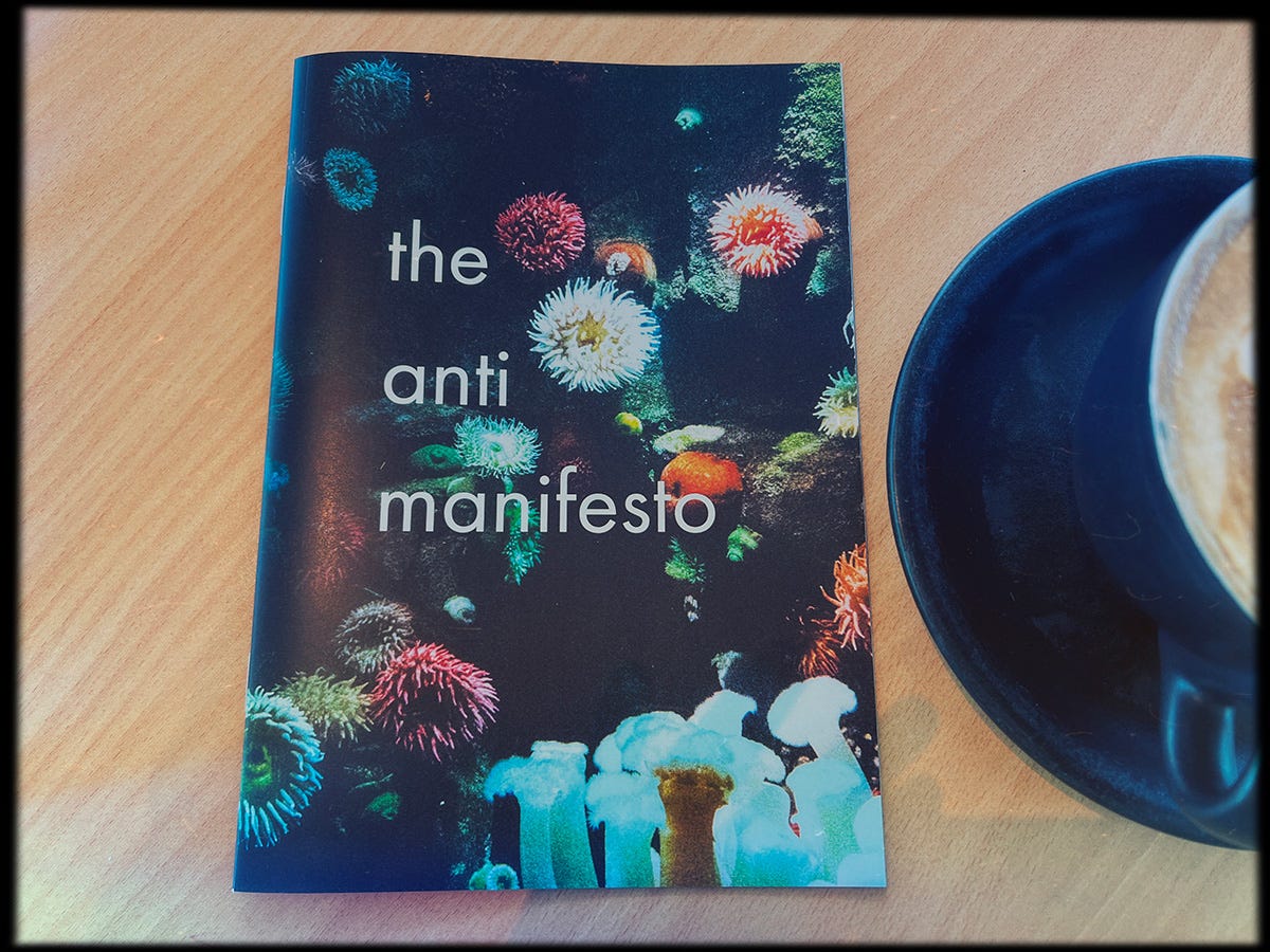 An A5 zine that has coral on the front cover with the words "the anti manifesto", it's next to a flat white coffee on a table