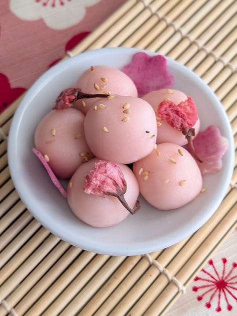 A trio of savory mochi in pink, white, and green, each plated with complementary garnishes. The pink mochi, infused with sakura powder, is topped with sesame seeds and pickled cherry blossoms. The white mochi, made from potatoes, is sprinkled with furikake and crispy fried onions. The green mochi, crafted from peas, is adorned with black sesame seeds and thinly sliced radish. Each piece highlights a unique texture and flavor, arranged for a visually striking presentation.