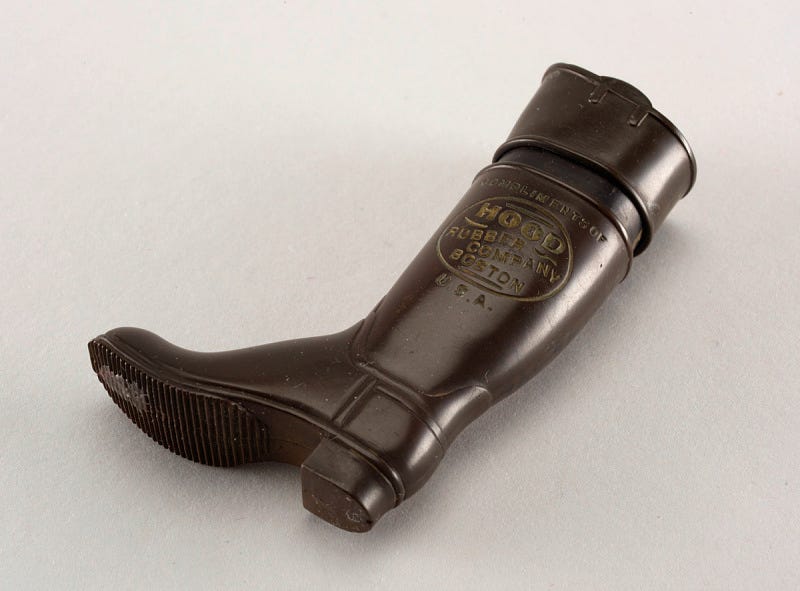 In the shape of a tall black boot, inscribed in light brown "Compliments of Hood Rubber Company, Boston, U.S.A.", with slip-top lid, which may be removed entirely. Striker on grooves of sole on boot bottom In the shape of a tall black boot, inscribed in light brown "Compliments of Hood Rubber Company, Boston, U.S.A.", with slip-top lid, which may be removed entirely. Striker on grooves of sole on boot bottom