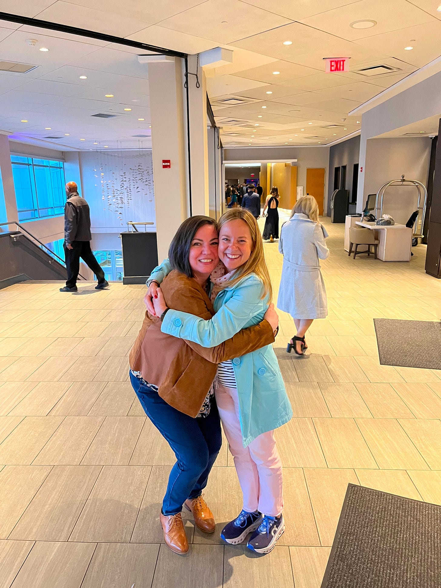 Two sisters hugging in a hotel lobby. One is taller and brunette with a short brown jacket, jeans, and brown shoes. The other is shorter, blonde, and wearing a long, light blue jacket, pink pants, and blue sneakers.