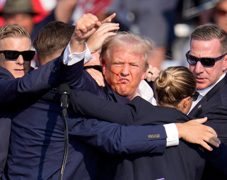 A man with greyish blond hair and a streak of blood on his face raises his fist in the middle of a huddle of Secret Service agents.