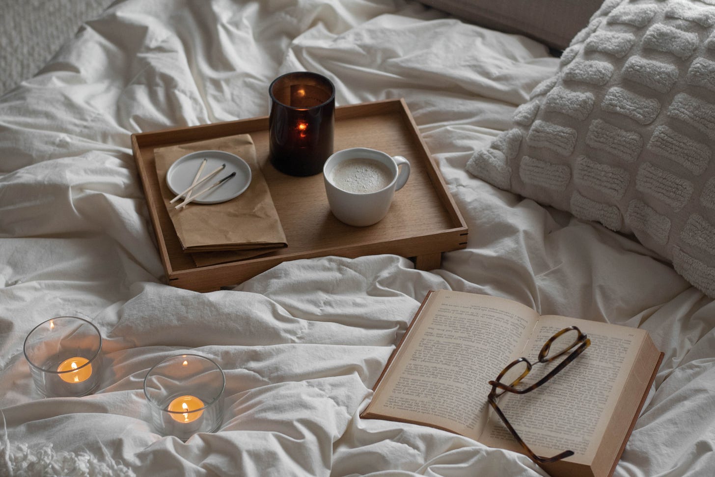 an open book lies on a rumpled bed, next to a tray with a tea and candles, the extinquished matches lying on a plate next to the tea