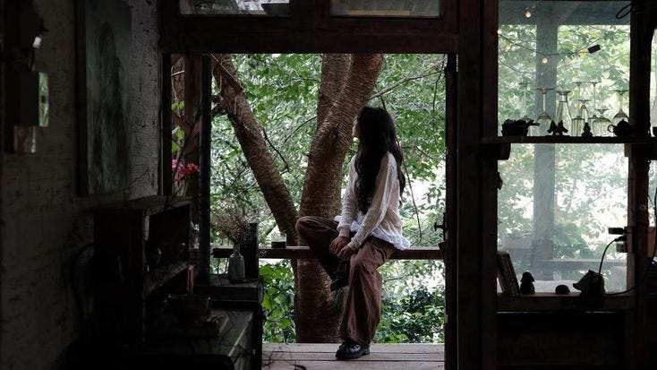 This may contain: a person sitting on a porch in front of a tree and looking out the window