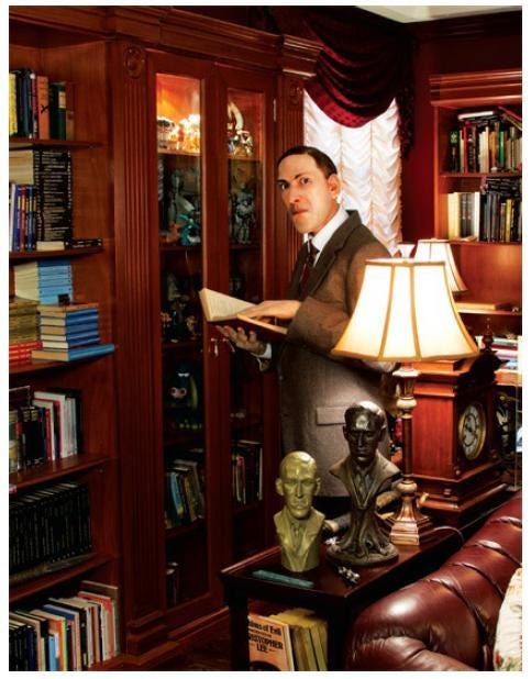 A life-sized statue of HP Lovecraft, a white man in a grayish suit, standing in among antique looking shelves full of books